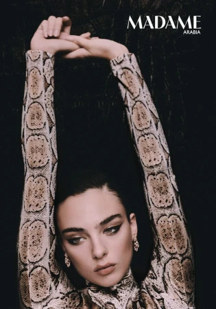 Model Deva Cassel posing with arms raised overhead wearing snakeskin print top against a dark background