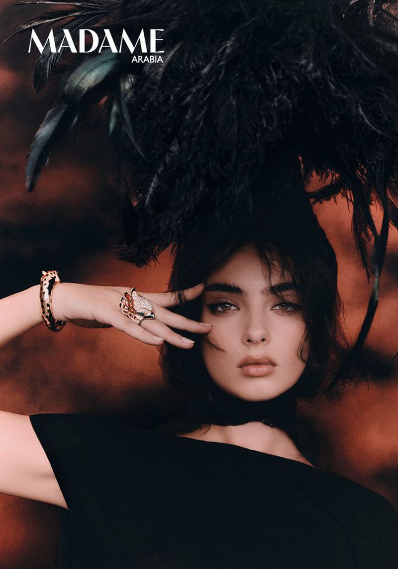 Deva Cassel wearing a black feathered hat and jewelry for a fashion editorial against a warm background.
