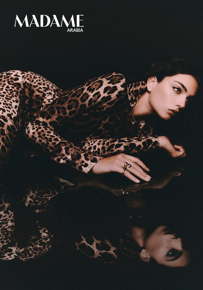 Deva Cassel reclining in a leopard print jumpsuit on a reflective surface against a dark background