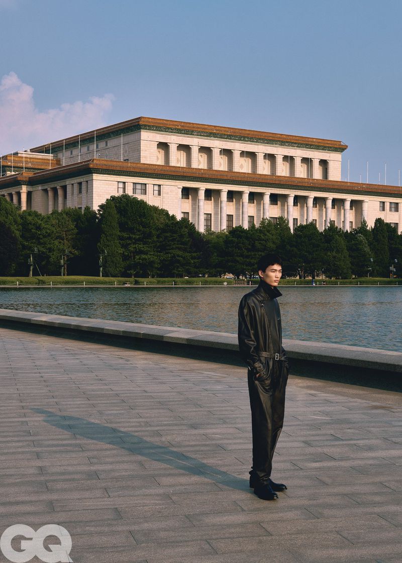 A man wearing a black leather jumpsuit standing on a paved plaza in front of a large neoclassical building and a body of water.