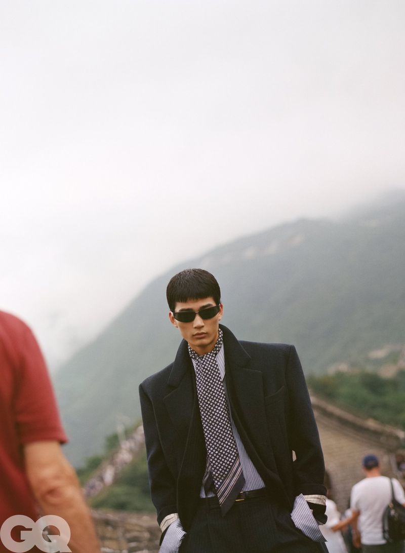 Man wearing dark sunglasses, patterned tie, and dark blazer outdoors against a mountain background with misty sky.