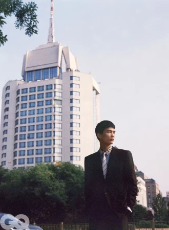 A man in a black suit and striped tie standing outdoors before a tall building in an editorial portrait.