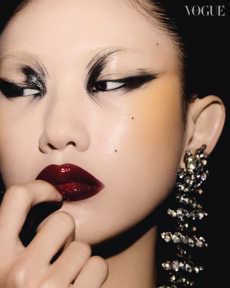 Close-up portrait of a model with dark dramatic eye makeup, glossy burgundy lips, and crystal chandelier earrings against a dark background.