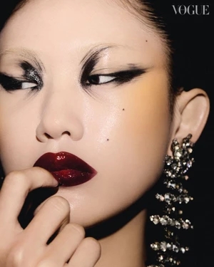 Close-up portrait of a model with dark dramatic eye makeup, glossy burgundy lips, and crystal chandelier earrings against a dark background.