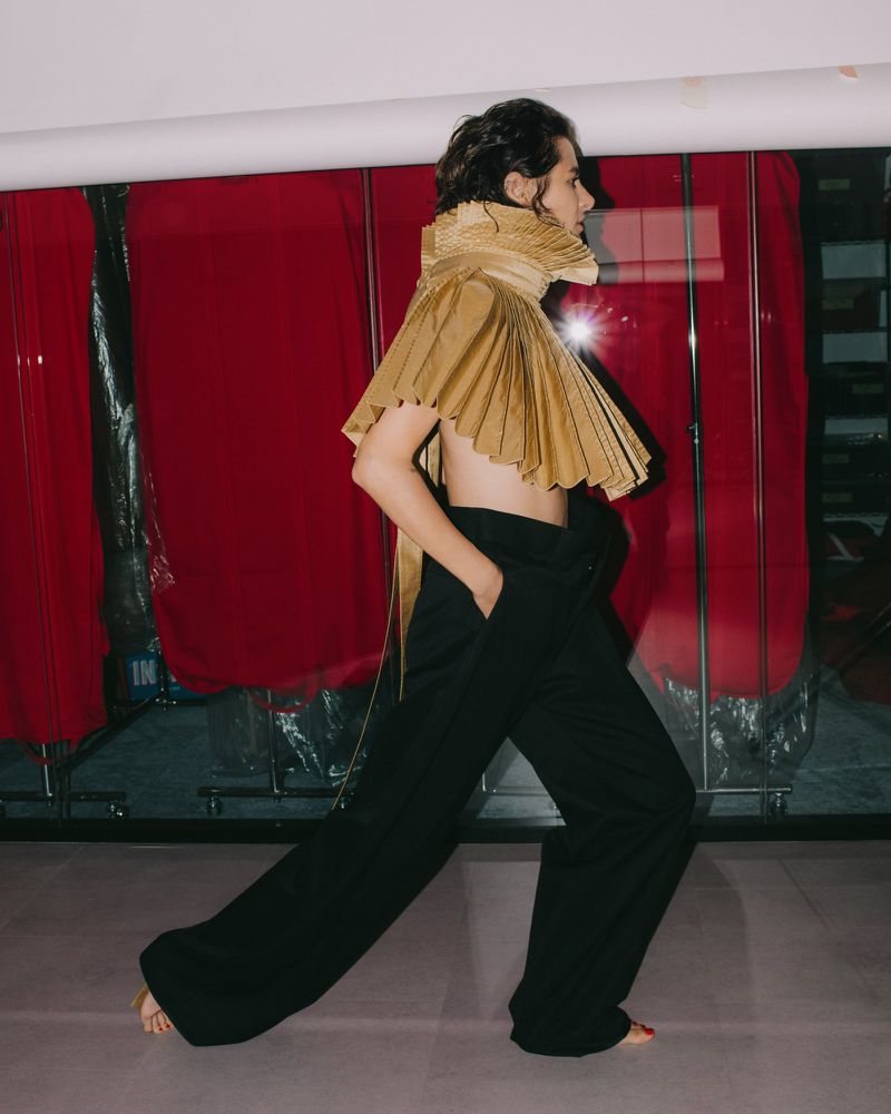 Model in profile wearing a pleated gold sculptural collar and wide black trousers inside a theatre setting