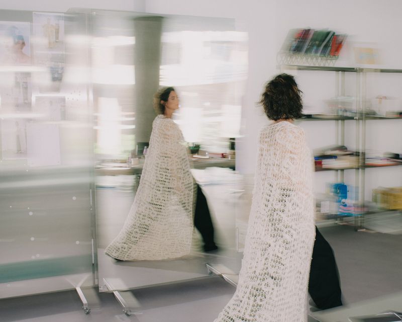A woman wearing a textured white knitted cape in an office setting with motion blur effect captured by Anastasiia Vlasova for i-D Magazine.