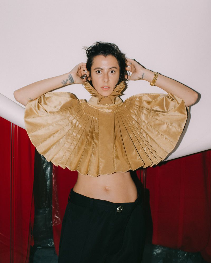 Woman wearing an elaborate pleated gold collar and matching cropped top with dark trousers against a white backdrop.