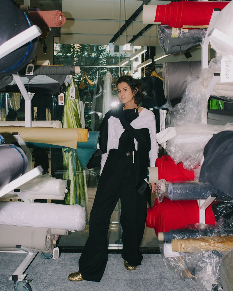 Woman standing amidst shelves stacked with rolls of fabric in a textile warehouse setting for i-D Magazine