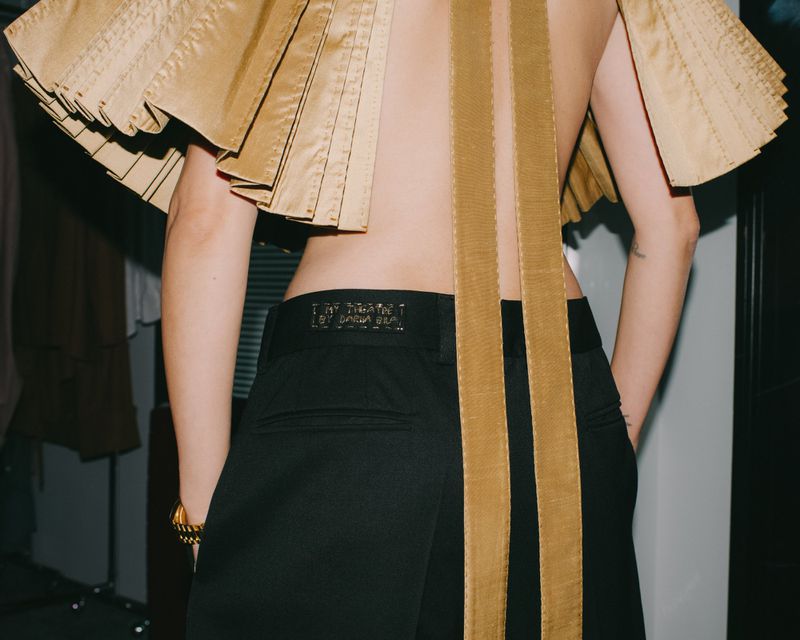 Model seen from the back wearing a pleated gold top and black trousers for i-D Magazine
