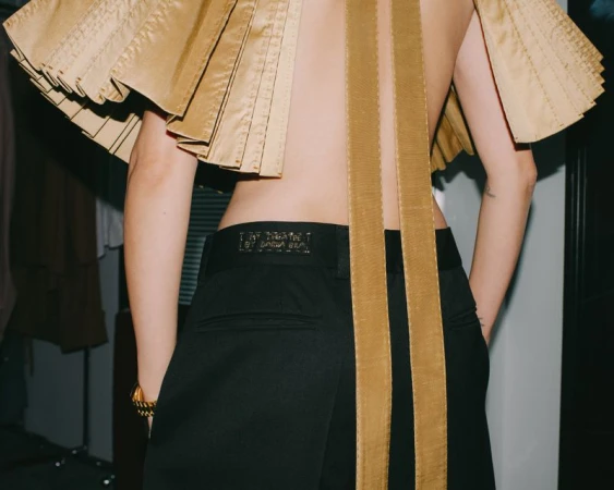 Model seen from the back wearing a pleated gold top and black trousers for i-D Magazine