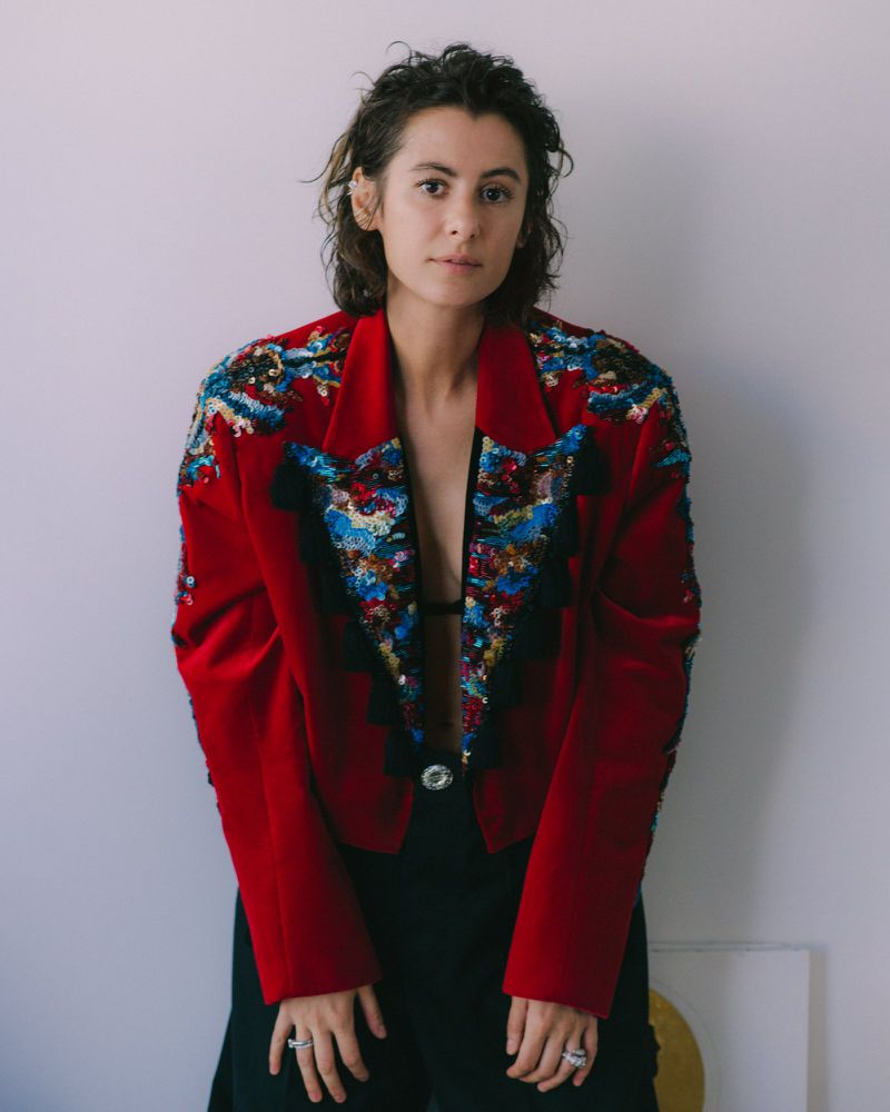 Portrait of a person with dark short hair wearing an embellished red blazer over dark trousers against a white seamless background.