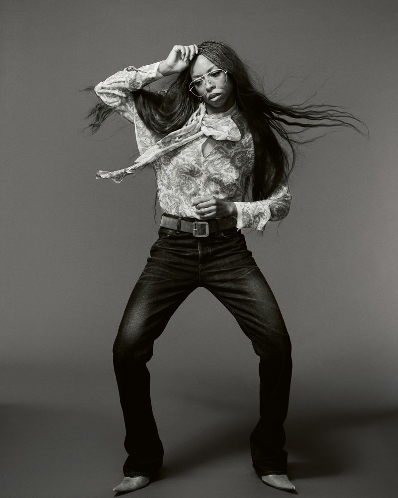Portrait of Ravyn Lenae in dark jeans and a patterned blouse with long dark hair blowing against a grey background.