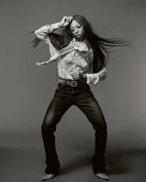 Portrait of Ravyn Lenae in dark jeans and a patterned blouse with long dark hair blowing against a grey background.