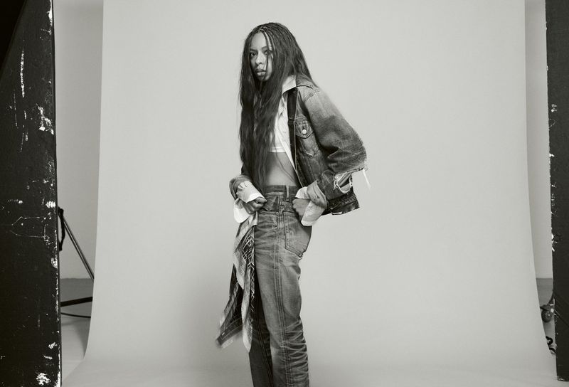 Portrait of Ravyn Lenae wearing denim jacket and jeans against a white seamless backdrop for Heroine Magazine.