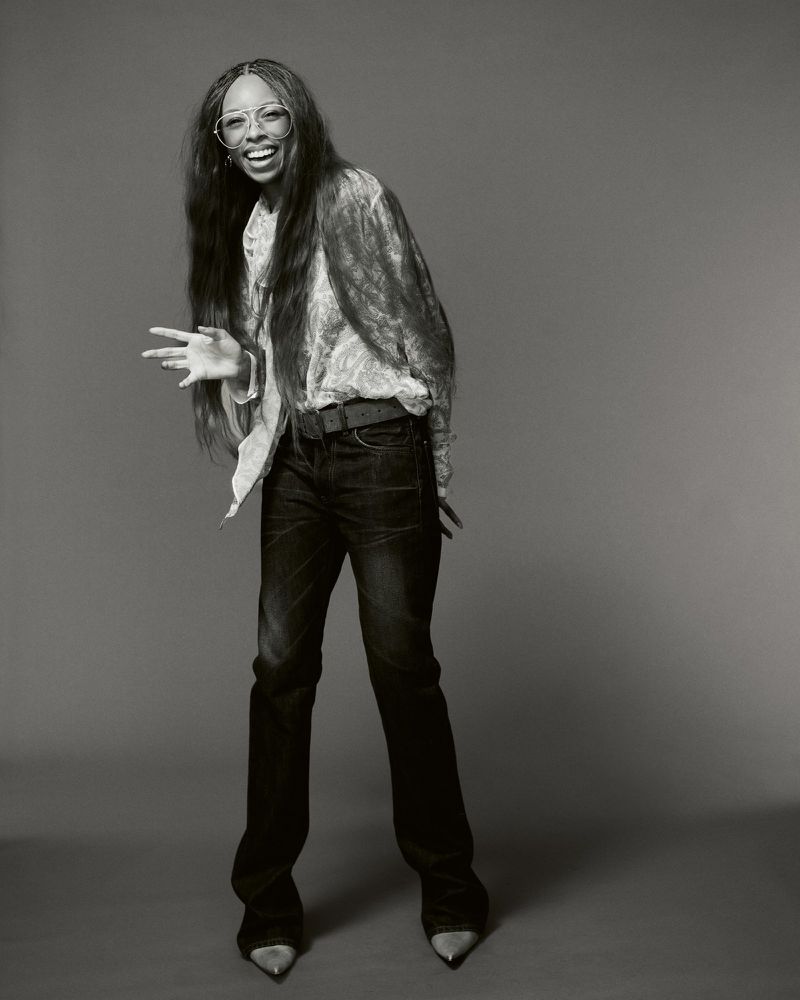Portrait of Ravyn Lenae smiling in dark jeans and a patterned blouse against a grey seamless studio backdrop for Heroine Magazine.