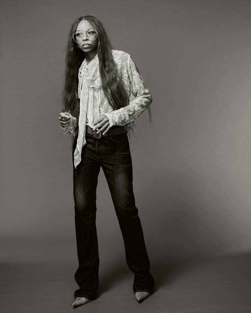 Portrait of Ravyn Lenae wearing a patterned blouse and dark trousers against a grey studio backdrop for Heroine Magazine.