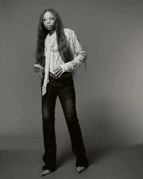 Portrait of Ravyn Lenae wearing a patterned blouse and dark trousers against a grey studio backdrop for Heroine Magazine.