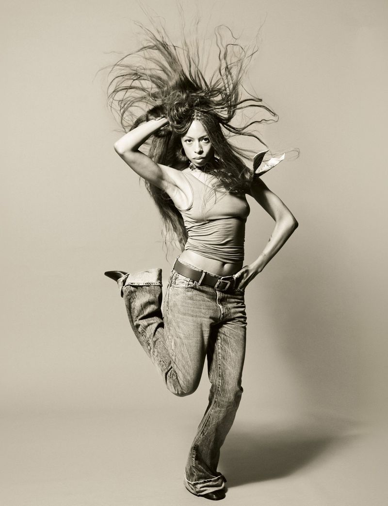 Portrait of Ravyn Lenae mid-jump with long hair flying, wearing a tank top and jeans against a plain studio background.