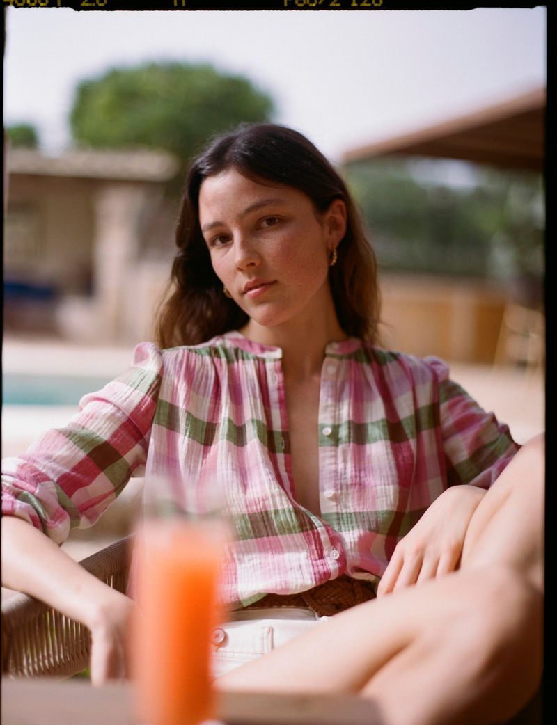 A young woman in a plaid shirt relaxes outside with a drink. Natural light highlights her features and the casual, summery setting.