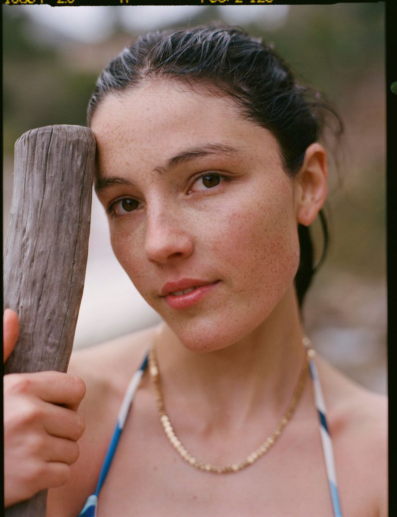 Portrait of a young woman with dark hair and freckles, wearing a blue bathing suit and gold necklace, standing outdoors near a weathered wooden post.