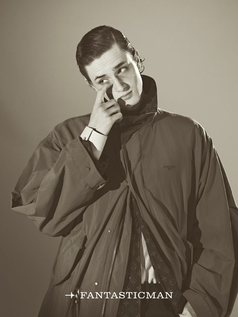 Monochrome portrait of a male model wearing a dark jacket with a high collar, wiping a tear from his eye against a light background.