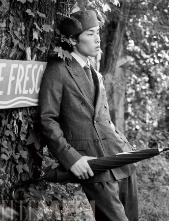 Monochrome fashion editorial featuring an East Asian male model leaning against a tree trunk wearing a tailored suit and cap.