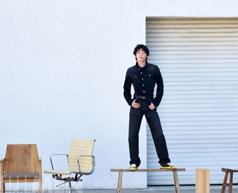 Model in dark denim jacket and pants standing on a wooden bench against a white wall with chairs in foreground
