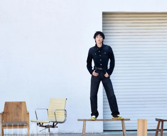 Model in dark denim jacket and pants standing on a wooden bench against a white wall with chairs in foreground