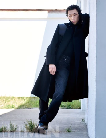 Model wearing a black coat and dark jeans leaning against a light-colored wall for Elle Men China