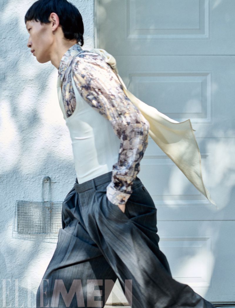 Profile of a male model in patterned shirt and grey trousers walking past a pale garage door for Elle Men China.