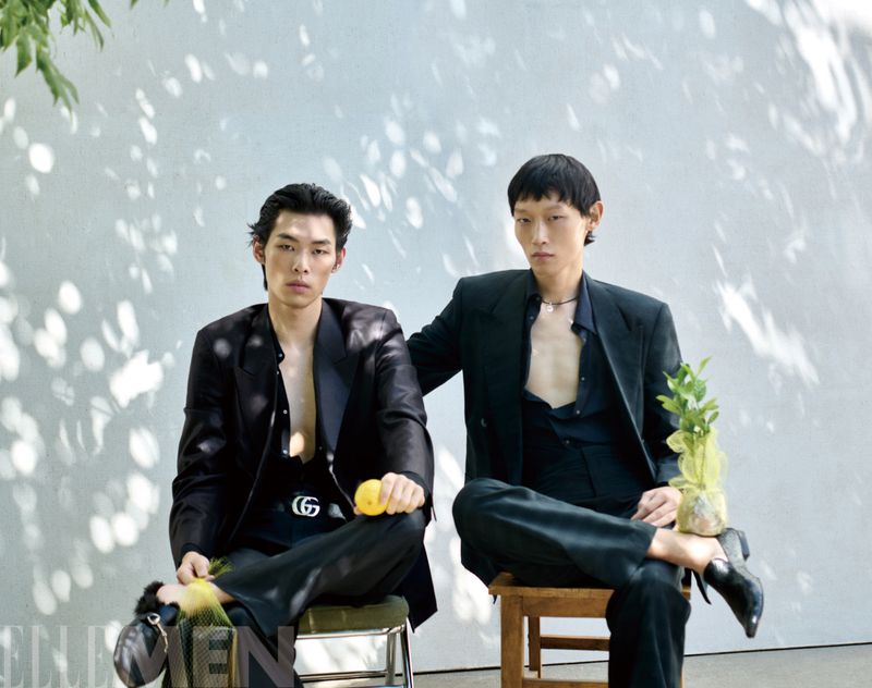 Two male models in dark suits seated outdoors against a pale wall with dappled sunlight shadows, holding citrus fruits.