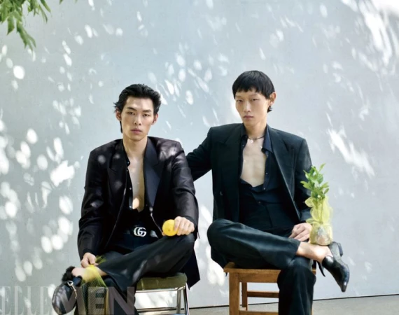 Two male models in dark suits seated outdoors against a pale wall with dappled sunlight shadows, holding citrus fruits.