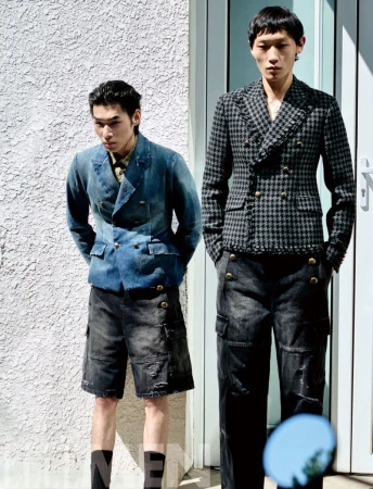 Two male models posing outdoors in front of a textured wall and doorway wearing structured double breasted jackets and dark trousers.