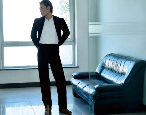 Man wearing a black suit and white shirt standing next to a leather sofa in an interior setting