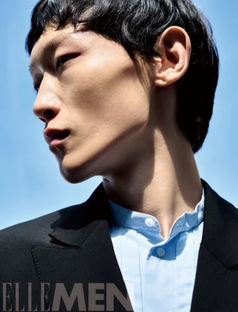 Close-up portrait of a male model wearing a black jacket and light blue shirt against a clear blue sky background.