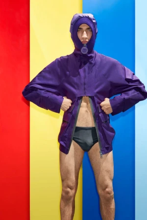 Man wearing a hooded purple jacket and dark briefs against a brightly colored vertical striped background.