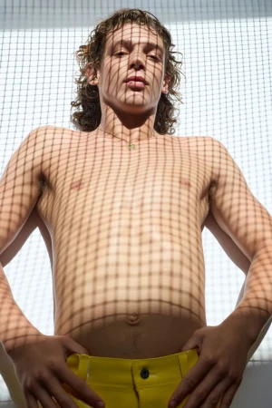 Shirtless young man with curly hair wearing yellow trousers against a white grid-patterned background under patterned light.