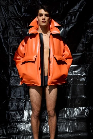 Male model wearing a bright orange jacket and dark briefs against a crinkled black plastic background.