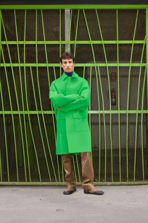 Man wearing a vibrant green coat and brown trousers standing before a green patterned security gate.