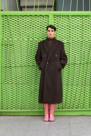 Man standing in a long brown coat and pink boots against a bright green patterned gate for Fantastic Man