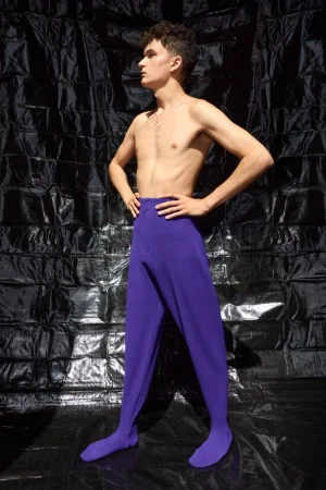Shirtless young man wearing purple pants standing against a crinkled black background