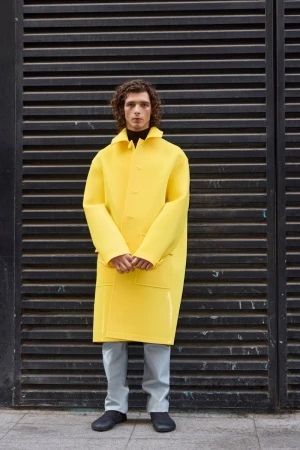Young man with curly hair wearing a bright yellow coat standing against dark slatted metal background