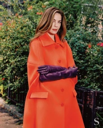 Michelle Monaghan in an orange coat and purple gloves posing before green foliage for S Magazine