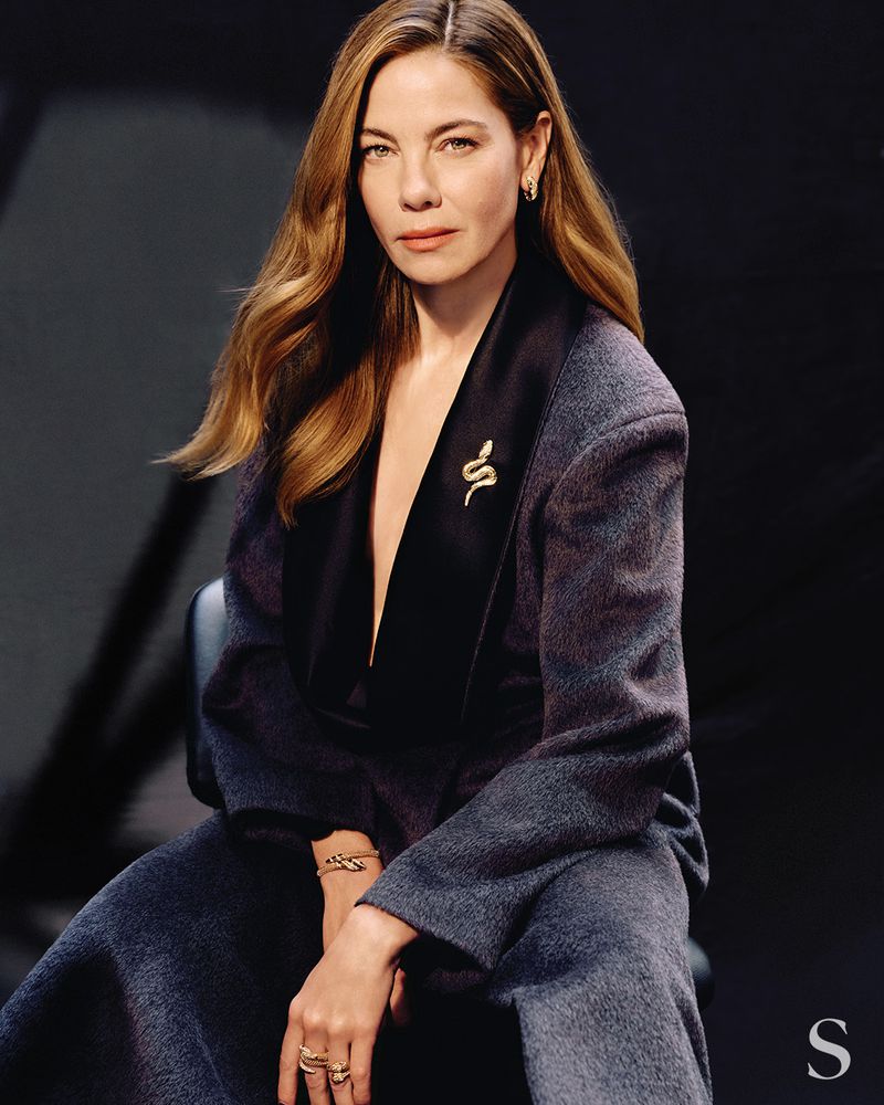 Michelle Monaghan seated wearing a dark textured blazer with satin lapel and gold snake jewelry against a dark background