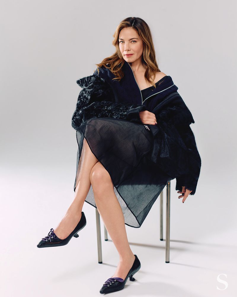 Michelle Monaghan seated on a metal chair in a dark textured jacket and sheer skirt against a white background
