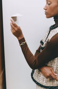 Woman in tweed vest holding a folded paper airplane against a plain white background for Monocle Magazine.
