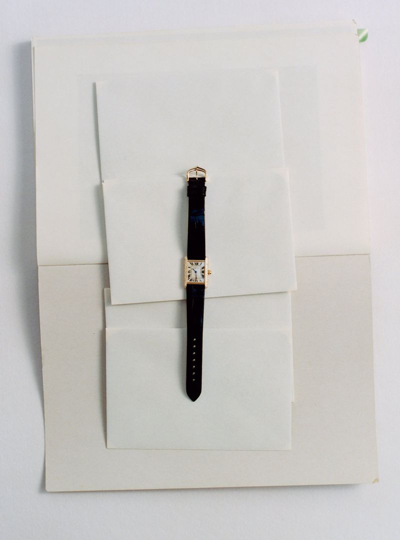 Gold wristwatch with black leather strap placed vertically across layered white envelopes and paper sheets.