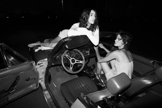 Two women posing around a classic convertible car at night in a high contrast black and white fashion editorial photograph.