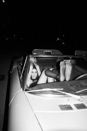 Black and white portrait of a woman reclining inside a convertible car at night with her feet up on the dashboard.