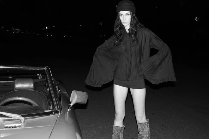 Black and white editorial portrait of a woman wearing a beanie and black flared sleeve top standing beside a vintage car at night.